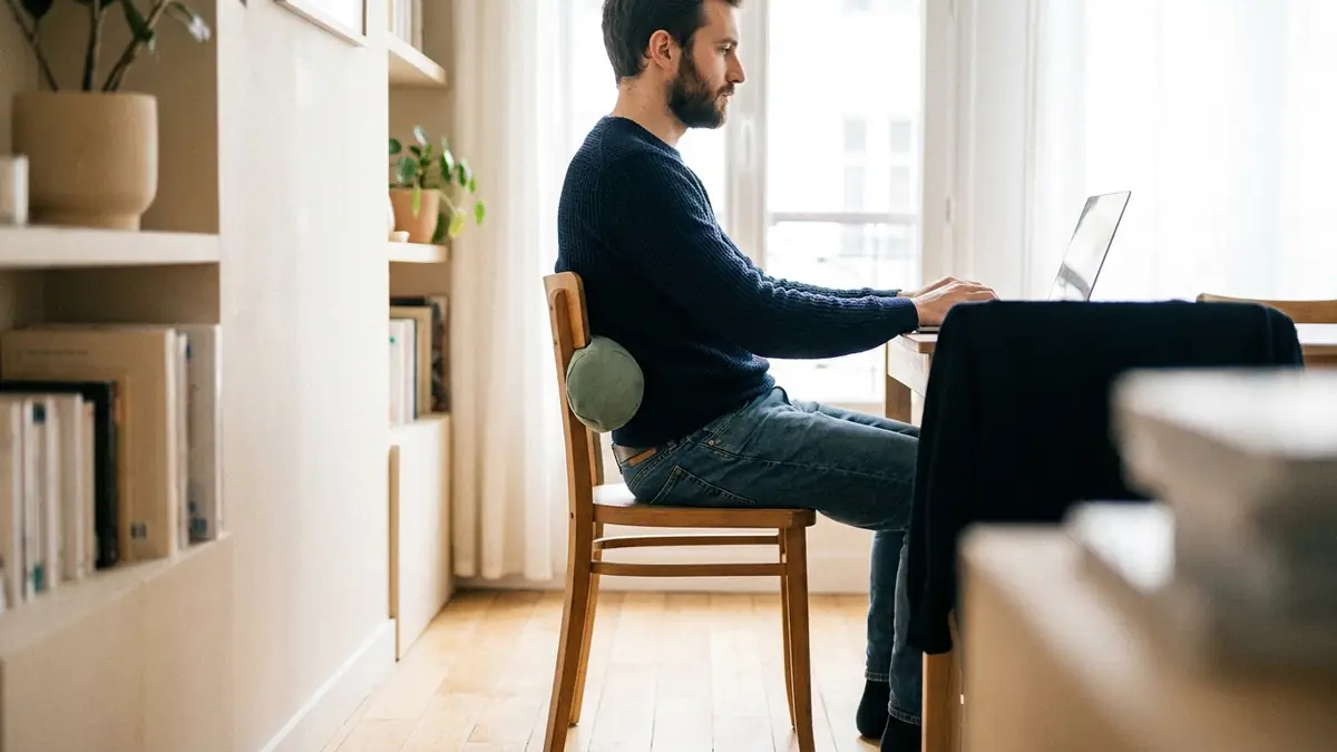 Homme en télétravail assis droit avec un coussin lombaire roulé dans le creux du dos, vu de profil