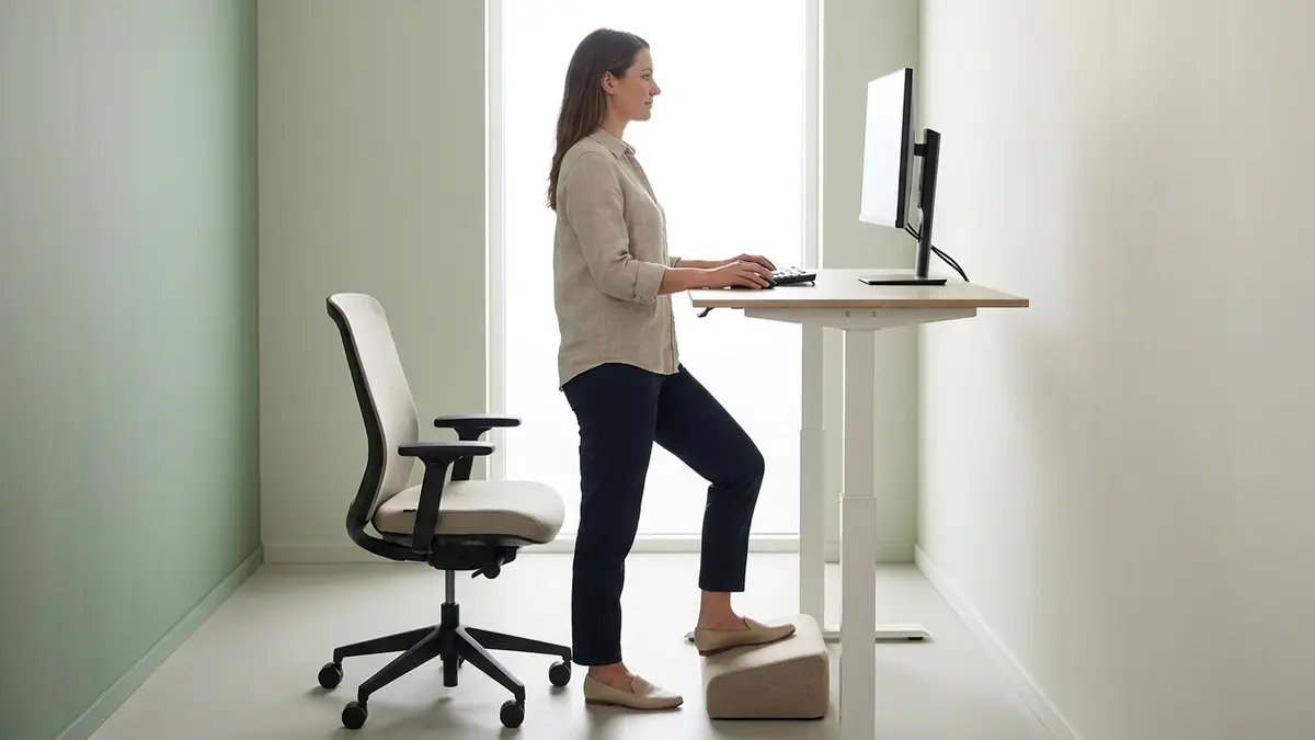 Femme qui travaille debout à un bureau réglable assis-debout, un pied sur un repose-pieds, sa chaise à côté pour alterner les postures